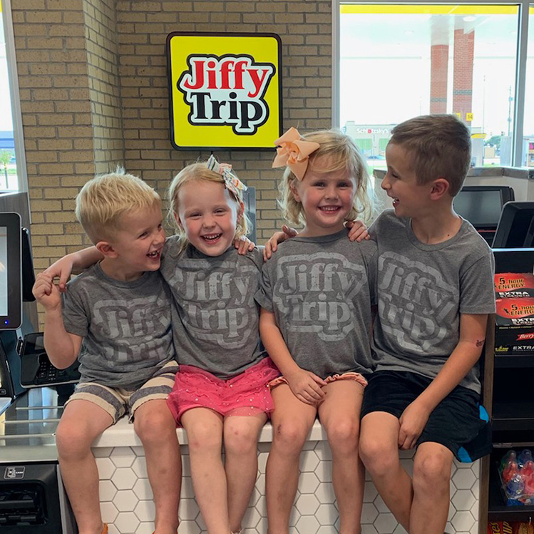 Four smiling Williams grandchildren in Jiffy Trip shirts sitting inside a Jiffy Trip store, showcasing the family-oriented nature of the company.